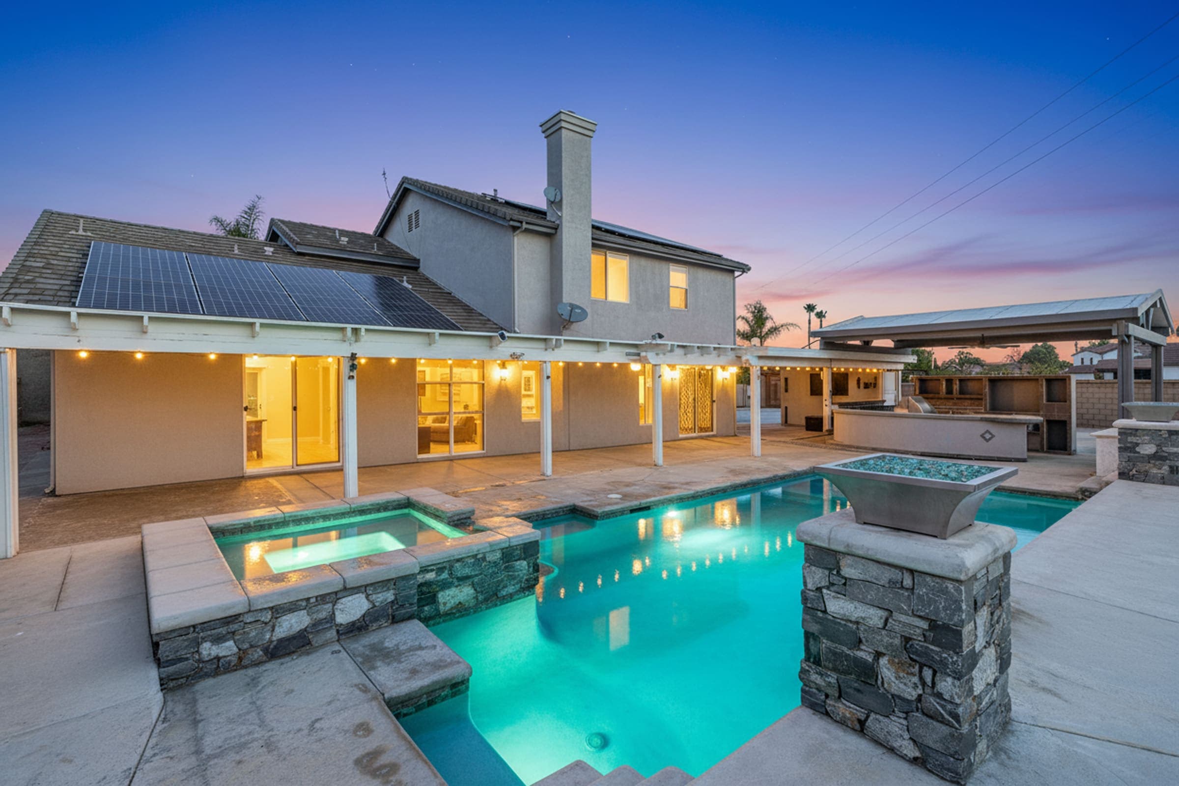 The pool, spa and covered patio behind a two-story Riverside home at dusk, lit against a blue and pink sky